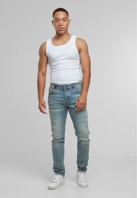 Giovane uomo con capelli corti indossa una maglietta bianca senza maniche, jeans blu strappati e sneaker bianche, in piedi davanti a uno sfondo grigio semplice.