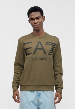 Mann trägt olivgrünes EA7 Emporio Armani Sweatshirt und hellblaue Jeans, steht vor einem einfarbigen grauen Hintergrund.
