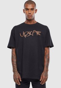 Sort bomulds T-shirt med et stort brunt grafitti-lignende logo på forsiden. Korte ærmer, rund hals, afslappet pasform og synlige tatoveringer.