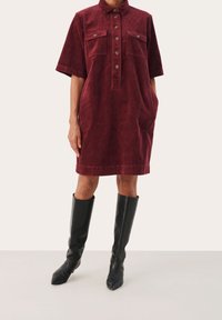 Burgunderrotes Cordkleid mit Kragen, kurzen Ärmeln, durchgehender Knopfleiste, zwei Brusttaschen und Seitentaschen, kombiniert mit schwarzen kniehohen Stiefeln.