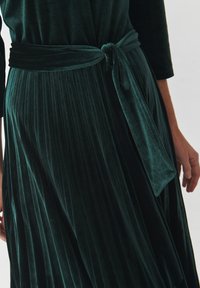 Emerald green velvet dress with pleated skirt. Features a tied waist sash and long sleeves, showcasing a soft, textured fabric.