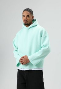 Sweat à capuche oversize vert menthe avec cordon de serrage à la capuche, manches longues et bord brut. Porté sur une chemise blanche et un pantalon noir. Texture lisse.