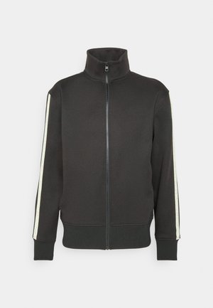 Giacca nera con zip e colletto alto, dotata di strisce laterali chiare che mostrano il nome del marchio. Realizzata in un tessuto liscio con polsini a costine.