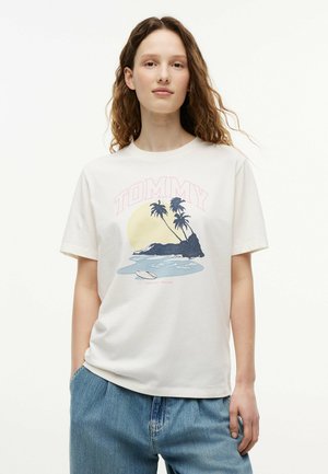 Młoda kobieta z falującymi włosami, ubrana w białą koszulkę z nadrukiem wyspy, palm, deski surfingowej, słońca i napisem "TOMMY", zestawioną z niebieskimi dżinsami.