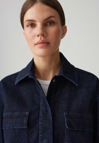 Giacca di jeans scura con colletto, due tasche sul petto e chiusura a bottoni. Presenta un tessuto texturizzato e una silhouette aderente.