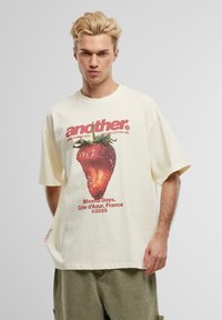 Cremefarbenes, oversized T-Shirt mit einem Grafikdesign einer Erdbeere, roten Texten und Designelementen. Aus Baumwolle, mit kurzen Ärmeln und lockerer Passform.