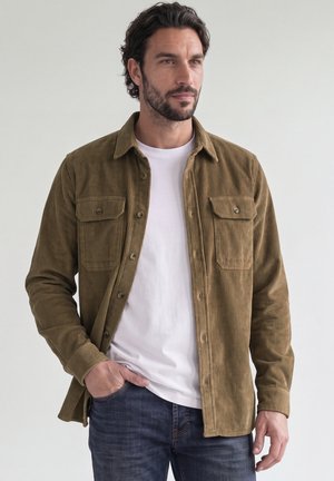 Uomo con capelli scuri e barba che indossa una camicia di velluto a coste marrone sopra una t-shirt bianca e jeans blu, in piedi davanti a uno sfondo uniforme.