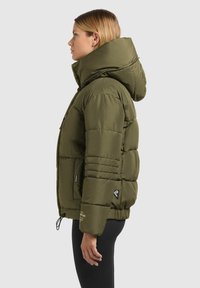 Veste matelassée vert olive avec un col montant, une grande capuche et des poches zippées. Présente une texture matelassée et un cordon de serrage à l'ourlet.