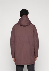 Parka bordeaux avec une capuche, dotée d'une texture lisse, d'une coupe droite et de coutures horizontales dans le dos, portée sur un pantalon noir.