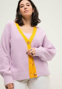 Cardigan lavorato a maglia viola con scollatura a V gialla profonda e chiusura con bottoni. Presenta polsini a costine e una vestibilità morbida, abbinato a un top leggero sottostante.