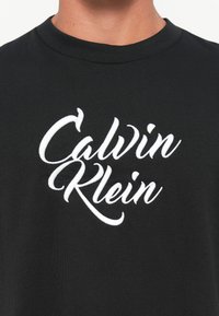 Camiseta negra de cuello redondo con el logo bordado en blanco "Calvin Klein" centrado en el pecho, usada por una persona.