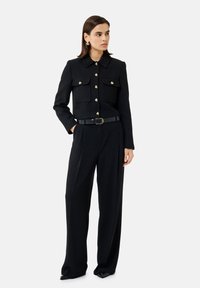 Blazer nera texturizzata con due risvolti sulle tasche e bottoni dorati, abbinato a pantaloni neri a gamba larga e una cintura nera. Presenti orecchini d'argento.