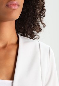 Blazer gris clair à texture lisse, avec un large col et des épaules structurées, partiellement visible sur l'épaule et le cou d'un modèle.