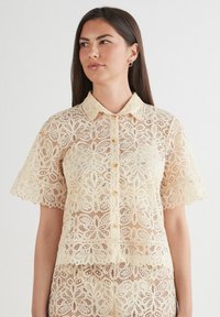Kantblouse in crèmekleur, met een kraag, korte mouwen en ingewikkelde bloemmotieven. Knopen aan de voorkant en een golvende zoom.