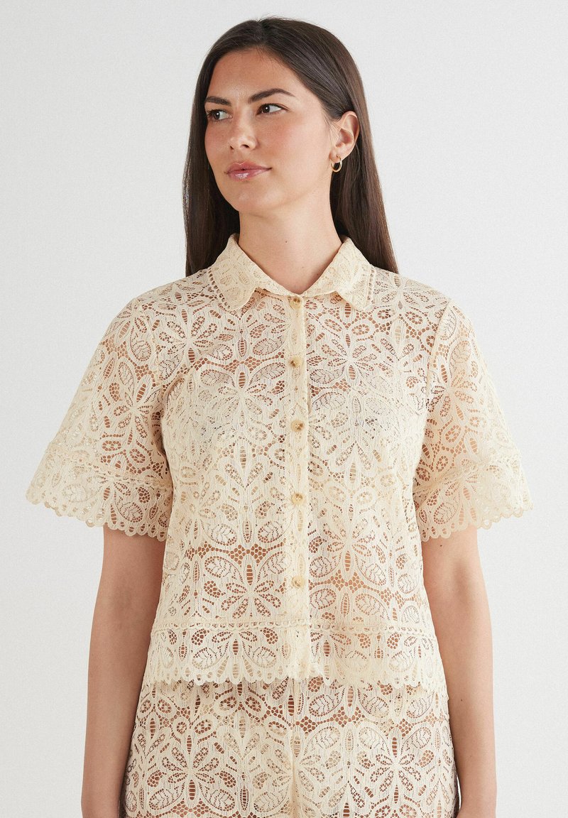 Kantblouse in crèmekleur, met een kraag, korte mouwen en ingewikkelde bloemmotieven. Knopen aan de voorkant en een golvende zoom.