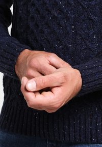 Mörkblå stickad tröja med en struktur av kabelmönster och ribbade muddärmar, bärs med hopknäppta händer och visar en subtil spräcklig detalj.