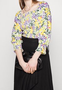Blouse à imprimé floral avec des citrons jaunes, des feuilles vertes et des fleurs violettes. Présente un design smocké, des manches longues et une coupe décontractée. Pochette noire.