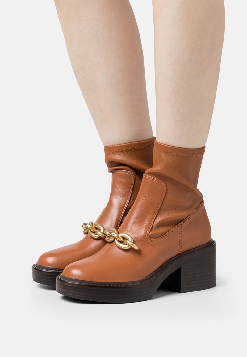 Bottines en cuir marron avec talons épais et grand détail de chaîne dorée portées sur des jambes nues sur fond blanc.