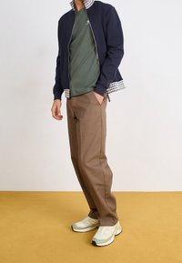 Giacca blu navy con zip e colletto a scacchi, t-shirt verde oliva, pantaloni beige e sneakers verde chiaro con dettagli grigi, su uno sfondo giallo.