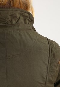 Zadní pohled na osobu oblečenou v olivově zelené bundě se šitými švy a viditelnou texturou látky na límci a rameni.