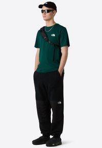 Zelené tričko s krátkým rukávem, černé fleecové kalhoty s lesklými černými akcenty, černá crossbody taška, sluneční brýle a černé pantofle.
