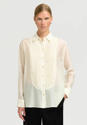 Lange mouwen, doorschijnende witte blouse met verticale plooien en een ronde zoom. Heeft een klassieke kraag en lichtgekleurde knopen.