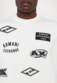 Biała bawełniana bluza z różnymi haftowanymi czarnymi logo i tekstami, w tym "Armani Exchange," "AX" oraz datą "1991."