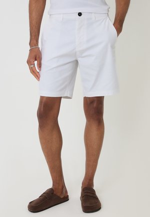Mand iført hvide shorts, hvid ærmeløs top, sølvarmbånd og ring samt brune slip-on loafers, stående med hænderne i lommerne.