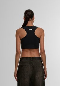 Black cropped tank top with racerback design. Made of smooth fabric, features white logo detail at the back. Paired with dark, textured pants.