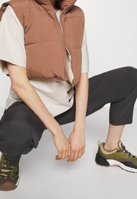 Person som sitter i huk med en beige kortärmad skjorta, brun pufferväst, svarta cropped byxor och gröna vandringsskor på en vit bakgrund.