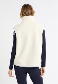 Kamizelka z polaru w kolorze kremowym z wysokim kołnierzem, ozdobiona niebieską lamówką i okrągłą naszywką z logo na ramieniu. Miękka, teksturowana powierzchnia.