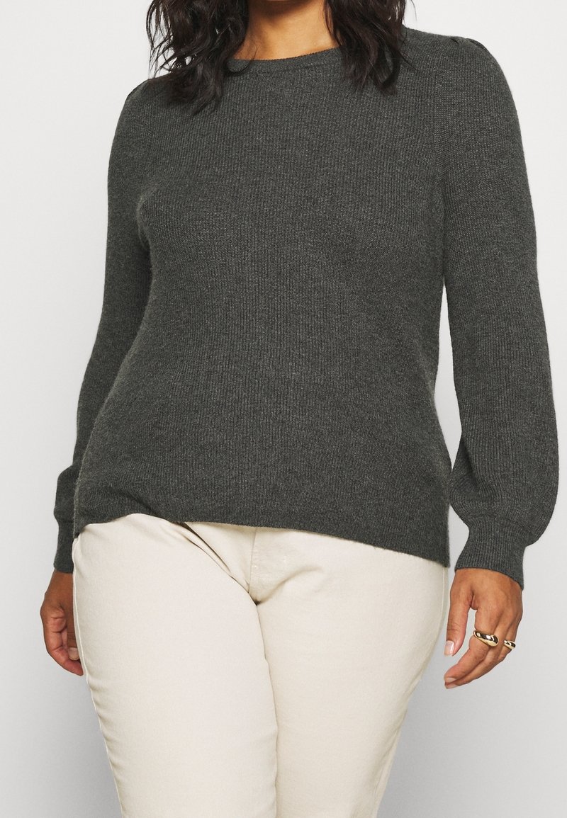 Ciemnoszary dzianinowy sweter z okrągłym dekoltem i lekko bufkowatymi długimi rękawami. Tekstura jest miękka i prążkowana. Noszony z beżowymi spodniami.