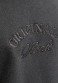 Szary materiał z wypukłym haftowanym napisem "ORIGINALS Studio" w dopasowanej ciemnej nitce.