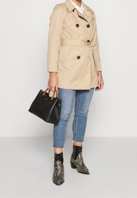 Trench-coat beige à boutons croisés et ceinture nouée. Jean bleu clair déchiré. Sac à main noir avec poignées en bois. Bottines en cuir de serpent.