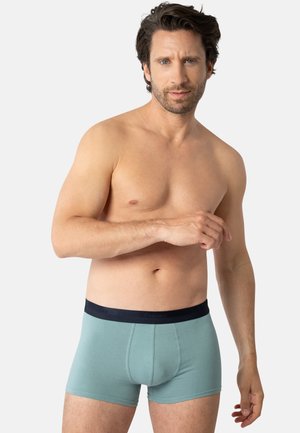 Boxer-briefs pour hommes en teal avec une ceinture navy. Tissu doux, design ajusté, et longueur mi-cuisse. Marquage minimal sur la ceinture.