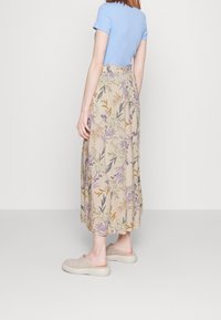 Jupe maxi florale beige avec des motifs violets, jaunes et verts ; taille élastique, associée à un haut ajusté bleu clair et des chaussures beiges à enfiler.