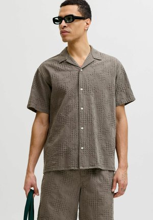 Mies pukeutunut kuvioituun, lyhythihaiseen nappipaitaan ja vastaaviin shortseihin, pitää tummaa vihreää laukkua, mustat aurinkolasit päässä, neutraali tausta.