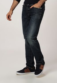 Tmavě modré džíny z denimového materiálu s slim fit střihem a jemným zesvětlením; kombinované s černými a hnědými teniskami s texturovaným povrchem a kontrastními detaily.