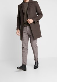 Manteau en laine marron avec un revers cranté, pantalon à carreaux gris et bottines noires. Le tissu est texturé, avec une coupe ajustée et un design structuré.