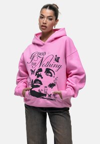 Felpa rosa brillante con vestibilità oversize, caratterizzata da una stampa grafica nera di un volto e farfalle. Include tasche a soffietto anteriori e cappuccio con cordino.