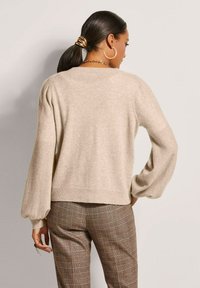 Beiger Strickpullover mit Ballonärmeln und geripptem Saum. Kombiniert mit braunen Karo-Hosen. Glatte Textur und lässige Passform.