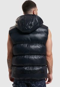 Schwarze Steppweste mit glänzender, gesteppter Oberfläche, ausgestattet mit einer Kapuze und einem Logo auf der Rückseite. Ärmelloses Design mit einer strukturierten Form.
