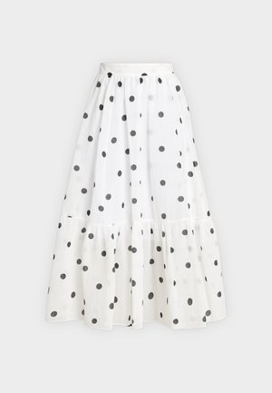 Femme portant un haut noir à manches courtes, une jupe blanche à pois noirs, des sandales noires à talons, tenant un sac à main blanc.