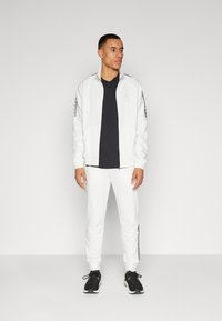 Giacca bianca con zip e dettagli neri, abbinata a pantaloni bianchi coordinati e scarpe da ginnastica nere. Il tessuto sembra liscio con un design pulito.