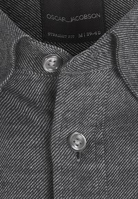 Gros plan sur le col d'une chemise grise à motif chevrons avec deux boutons et une étiquette indiquant Oscar Jacobson, coupe droite, taille M/39-40.