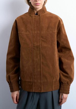 Vrouw draagt een bruine suède jas met geplooide manchetten en een hoge kraag, gecombineerd met donkergrijze broek tegen een effen witte achtergrond.