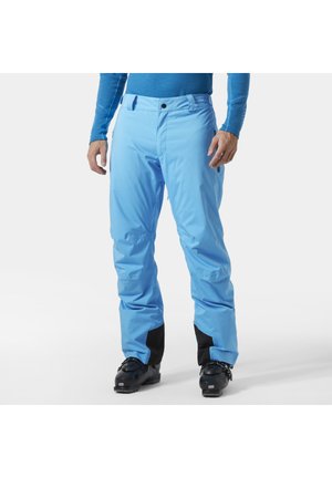 Mann trägt leuchtend blaue wasserdichte Hose mit schwarzen Skischuhen, steht vor weißem Hintergrund.