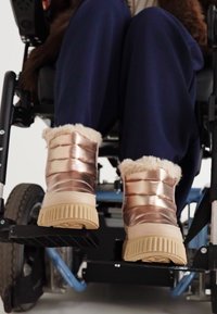 Bottes chelsea en or rose, rembourrées, avec une doublure en fourrure, une semelle en caoutchouc beige et des nervures texturées, présentées sur un repose-pieds de fauteuil roulant.