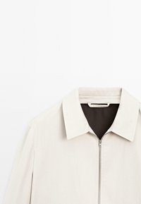 Lys beige zip-jakke med en struktureret krave, der har en glat stoftekstur og et kontrasterende mørkt indre.