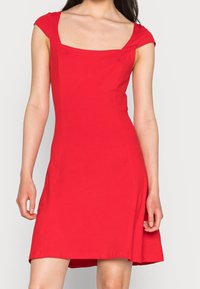 Mujer vestida con un vestido rojo brillante entallado, con mangas cortas y escote cuadrado, de pie frente a un fondo claro y liso.